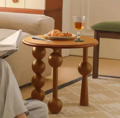 Ronde houten design bijzettafel met bolvormige poten – tafelblad met croissants, glas thee en pen, geplaatst in gezellige woonkamer met beige bank, groene stoel en persoon die leest.