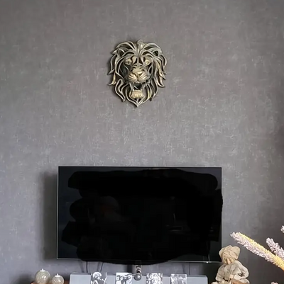 Wandsculptuur van leeuwenkop in glanzend goudkleurige kunsthars boven televisie op grijze gestructureerde muur in moderne woonkamer; toont realistische manen, expressieve blik en open bek, omringd door decoraties zoals glazen beeldjes, vaas met droogbloemen en een kindfiguur, een krachtig en elegant accent in het interieur