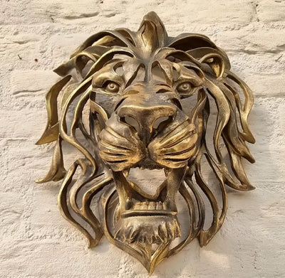 Wandsculptuur van leeuwenkop in glanzend goudkleurige kunsthars, gemonteerd op witte bakstenen muur; toont realistische manen en expressieve blik met open bek en zichtbare tanden, een sierlijk en imposant decoratief accent voor binnen- of buitengebruik