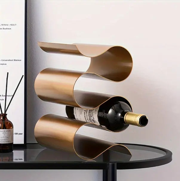 rijstaand wijnrek in koperkleurig metalen design met drie golvende houders, gepresenteerd op ronde glazen tafel met zwart frame, naast bruin diffuserflesje met geurstokjes; moderne en decoratieve presentatie in stijlvol interieur met witte muur en minimalistische kunst