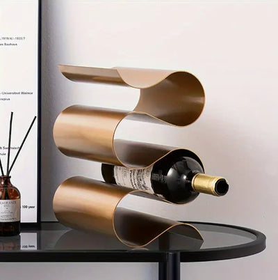 rijstaand wijnrek in koperkleurig metalen design met drie golvende houders, gepresenteerd op ronde glazen tafel met zwart frame, naast bruin diffuserflesje met geurstokjes; moderne en decoratieve presentatie in stijlvol interieur met witte muur en minimalistische kunst