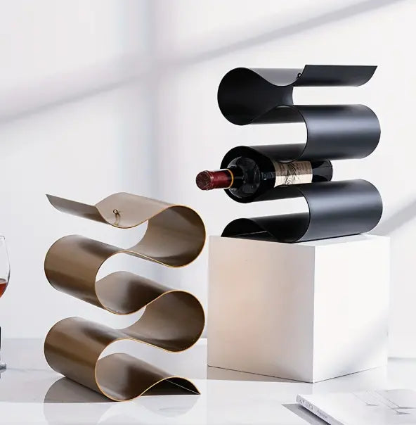 Twee vrijstaande wijnrekken met golvend metalen design, één in zwart en één in goudkleurig afgewerkt metaal, gepresenteerd met wijnfles en glas in een moderne setting; combineren creatieve vormgeving met functionele opslag voor decoratieve toepassing in keuken, bar of woonkamer