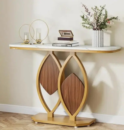 Console tafel met wit marmeren blad en sculpturaal onderstel in goudkleurig metaal, bestaande uit twee ovale ringen met houten diamantvormige inleg; decoratief gestyled met glazen kandelaars, houten doosje, boeken en vaas met groen-rode bladeren, geplaatst tegen lichte muur op visgraatvloer