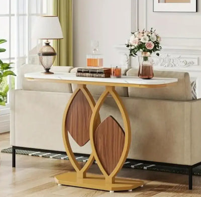 Console tafel met wit marmer-look blad en sculpturaal onderstel in goudkleurig metaal en houten diamantaccenten, geplaatst achter een beige bank in een moderne woonkamer met witgelambriseerde muur, groene gordijnen en decoratieve elementen zoals lamp, bloemenvaas, boeken en oranje glazen karaf