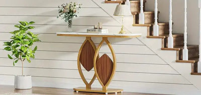 Console tafel met wit marmer-look blad en sculpturaal onderstel in goudkleurig metaal met houten diamantaccenten, gedecoreerd met vaas met bloemen, boekenstapel, lamp en kaars; geplaatst tegen witte lambrisering met houten trap en groene kamerplant in een eigentijds interieur