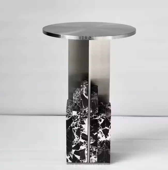 Moderne bijzettafel met rond metalen tafelblad en contrasterende voet in gepolijste staal en zwart marmer met witte aders, sculpturaal vormgegeven en visueel krachtig als interieurstatement