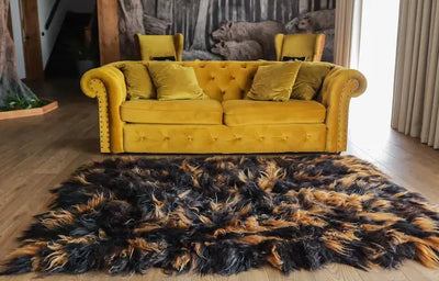 Fluweelzachte dierenhuid in zwart- en bruintinten, nonchalant gedrapeerd over een lichtgekleurde houten vloer voor een mosterdgele fluwelen sofa in rustieke woonkamer met boswandtapijt en natuurlijke accenten