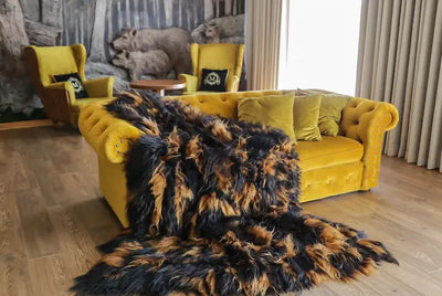 Grote dierenhuid met glanzende, langharige vacht in zwart- en bruintinten, losjes gedrapeerd over een okergele fluwelen sofa, als warm accent in een sfeervolle woonkamer