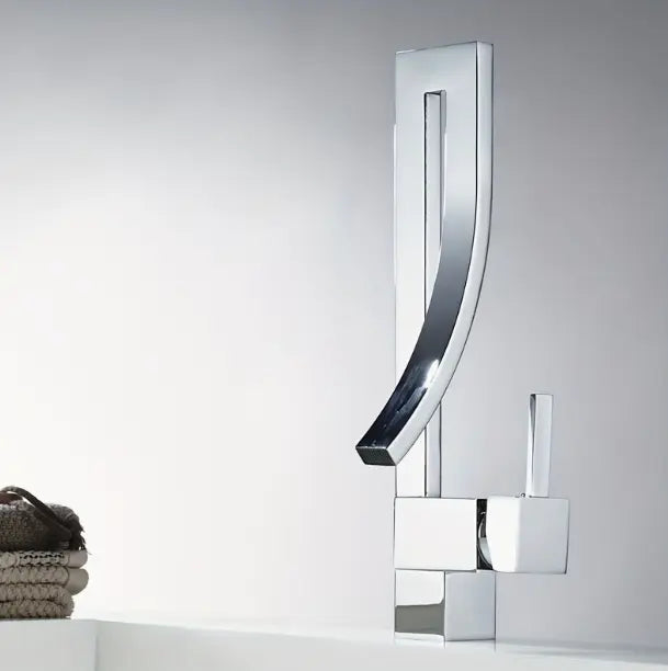 Design wastafelkraan in glanzend chroom met gebogen uitloop en eengreeps bediening, gemonteerd op witte wastafel met stapel opgerolde handdoeken ernaast; elegant accent in een moderne badkamer met neutrale achtergrond