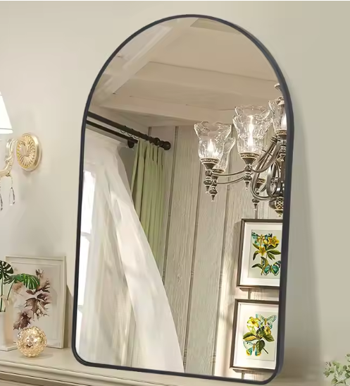 Boogvormige wandspiegel met zwarte metalen rand, gemonteerd op muur boven beige oppervlak met decoratieve elementen zoals een kleine plant en ingelijst kunstwerk; in de spiegelreflectie zijn een kroonluchter met glazen lampen, een witte gordijnroede en houten wandpanelen zichtbaar, evenals twee florale schilderijen; een elegant en ruimtelijk design dat klassieke charme combineert met moderne finesse voor hal, slaapkamer of woonkamer