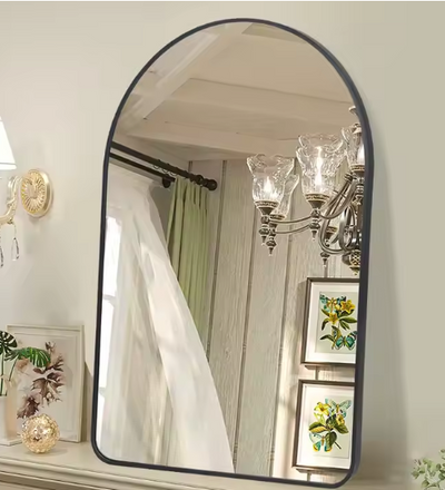 Boogvormige wandspiegel met zwarte metalen rand, gemonteerd op muur boven beige oppervlak met decoratieve elementen zoals een kleine plant en ingelijst kunstwerk; in de spiegelreflectie zijn een kroonluchter met glazen lampen, een witte gordijnroede en houten wandpanelen zichtbaar, evenals twee florale schilderijen; een elegant en ruimtelijk design dat klassieke charme combineert met moderne finesse voor hal, slaapkamer of woonkamer
