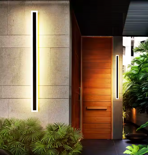 Moderne buitenlampen naast houten voordeur – verticale LED wandverlichting met warme gloed tegen lichtstenen gevel, stijlvolle entree met groene planten en designmaterialen.