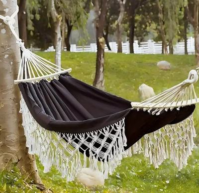Luxe Braziliaanse hangmat in diep zwart met witte macramé franjes, opgehangen tussen twee bomen in een groene tuin. De hangmat heeft een spreidstok aan beide uiteinden en hangt boven gras, met een witte omheining en rotsen op de achtergrond — een serene en stijlvolle plek voor ontspanning in bohemien sfeer.