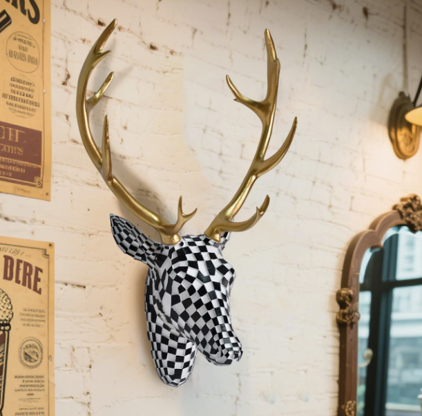 Wanddecoratie van hertenkop in zwart-wit geblokt mozaïekpatroon met glanzend gouden gewei, gemonteerd op witte bakstenen muur met links poster met tekst en rechts decoratieve spiegel; opvallend en artistiek accent voor modern interieur met creatieve flair