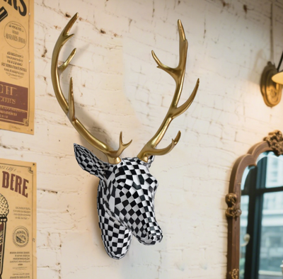 Wanddecoratie van hertenkop in zwart-wit geblokt mozaïekpatroon met glanzend gouden gewei, gemonteerd op witte bakstenen muur met links poster met tekst en rechts decoratieve spiegel; opvallend en artistiek accent voor modern interieur met creatieve flair