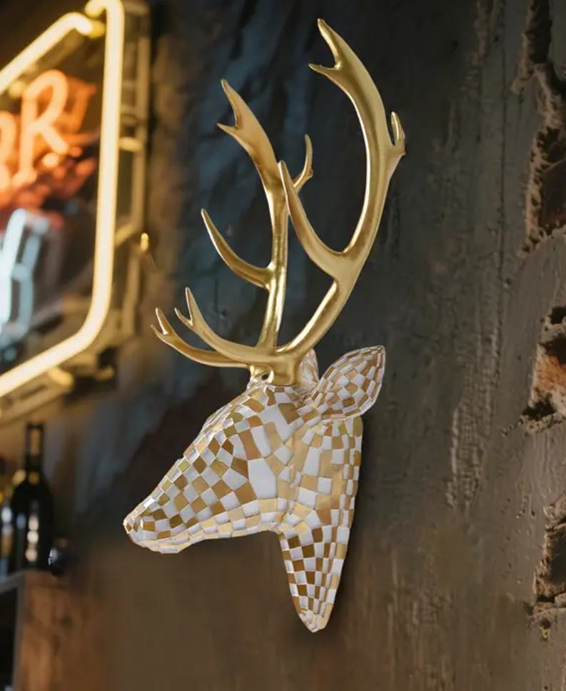 Wanddecoratie van hertenkop in zwart-wit geblokt mozaïekpatroon met glanzend gouden gewei, gemonteerd op witte gestructureerde muur met daarachter neonlicht en wijnfles op decoratief wandrek; artistieke combinatie van natuurmotief en modern design in eigentijdse setting