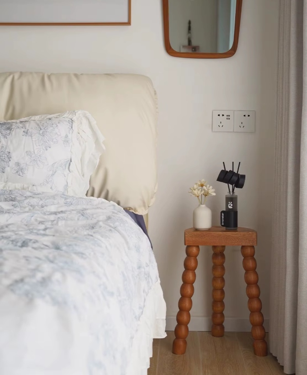 Gezellige slaapkamerhoek met een bed in wit-blauw bloemenpatroon en een houten bijzettafel met bolvormige poten. De tafel is gestyled met een vaas met witte bloemen, een zwart smart home-apparaat en een afstandsbediening. Op de muur erboven hangen twee spiegels en stopcontacten — een serene en functionele ruimte met natuurlijke tinten en sculpturaal design.