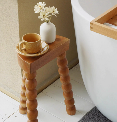 Luxe houten bijzettafel met sculpturale bolpoten, geplaatst naast een witte badkuip op wit houten vloer. De tafel toont een speels en artistiek ontwerp met drie beaded poten en is gestyled met een witte vaas met bloemen en een gele kop en schotel — een elegant accent voor badkamer, slaapkamer of woonkamer.