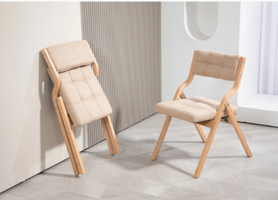 Twee opklapbare houten stoelen met beige gewatteerde zit- en rugkussens in tufted patroon; één stoel uitgeklapt en geplaatst op lichte vloer, de andere opgevouwen tegen witte wand, binnen setting met minimalistische inrichting; een boheemse en praktische zitoplossing voor interieur, evenement of kleine ruimte