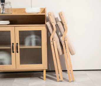 Interieurafbeelding met houten vitrinekast met glazen deuren, gevuld met borden en kommen, en twee houten opklapbare stoelen zonder kussens, schuin tegen een witte muur geplaatst; kast gedecoreerd met glazen potten en schalen bovenop, vloer bekleed met lichte tegels; een rustieke en functionele opstelling voor keuken, eetkamer of evenement met boheemse sfeer
