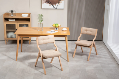 Sfeervolle interieurafbeelding met houten eettafel en twee opklapbare stoelen voorzien van beige gewatteerde zit- en rugkussens in tufted patroon; tafel gedecoreerd met fruit, kopje en plantje, geplaatst op lichtgrijze visgraatvloer; achtergrond toont houten sideboard met glazen deuren en abstracte wandkunst, een stijlvolle en praktische setting voor keuken, eetkamer of evenement