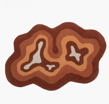 Fluffy vloerkleed in organische vorm met contourachtig patroon in beige, oranje en bruine tinten; ontwerp doet denken aan topografische lijnen of abstracte eilandstructuur met lichtere kleuren centraal en donkere aan de randen; afgebeeld zonder meubilair op neutrale achtergrond, een zacht en hoogpolig tapijt dat warmte en karakter toevoegt aan modern interieur