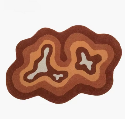 Fluffy vloerkleed in organische vorm met contourachtig patroon in beige, oranje en bruine tinten; ontwerp doet denken aan topografische lijnen of abstracte eilandstructuur met lichtere kleuren centraal en donkere aan de randen; afgebeeld zonder meubilair op neutrale achtergrond, een zacht en hoogpolig tapijt dat warmte en karakter toevoegt aan modern interieur