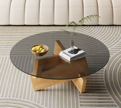 Moderne ronde salontafel in Art Deco-stijl met transparant gehard glasblad en sculpturaal houten onderstel; tafel gedecoreerd met gele fruitschaal, CHANEL-boek en glazen vaas met groene varen, geplaatst op grafisch vloerkleed met golvend patroon, naast beige gestoffeerde sofa, een stijlvol en luchtig interieuraccent voor eigentijdse woonruimte
