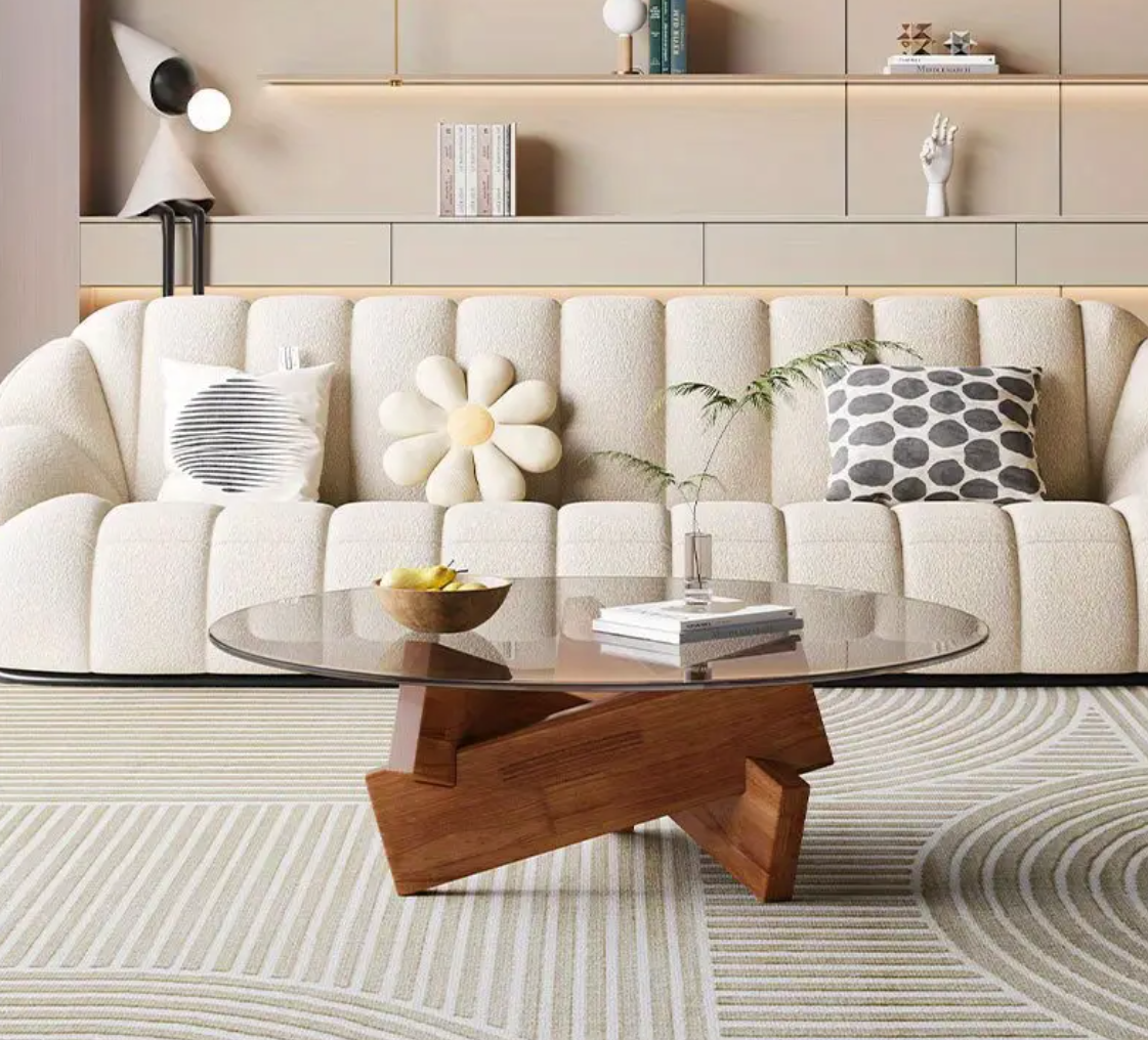 Moderne ronde salontafel in Art Deco-stijl met transparant gehard glasblad en stevig houten frame in natuurlijke houtkleur; geplaatst op grafisch patroonkleed voor beige sofa met drie decoratieve kussens, waaronder bloemvorm en gestreept patroon; tafel gedecoreerd met glazen vaas met plant, gele schaal en boek, omringd door ingebouwde wandrekken met decoratieve objecten, een elegant en luchtig middelpunt in stijlvol interieur