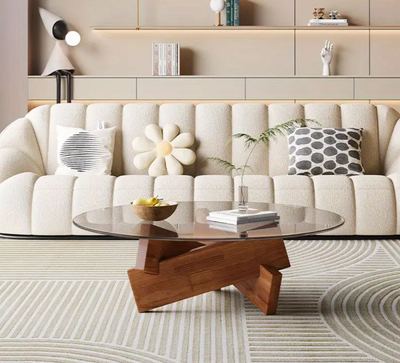 Moderne ronde salontafel in Art Deco-stijl met transparant gehard glasblad en stevig houten frame in natuurlijke houtkleur; geplaatst op grafisch patroonkleed voor beige sofa met drie decoratieve kussens, waaronder bloemvorm en gestreept patroon; tafel gedecoreerd met glazen vaas met plant, gele schaal en boek, omringd door ingebouwde wandrekken met decoratieve objecten, een elegant en luchtig middelpunt in stijlvol interieur