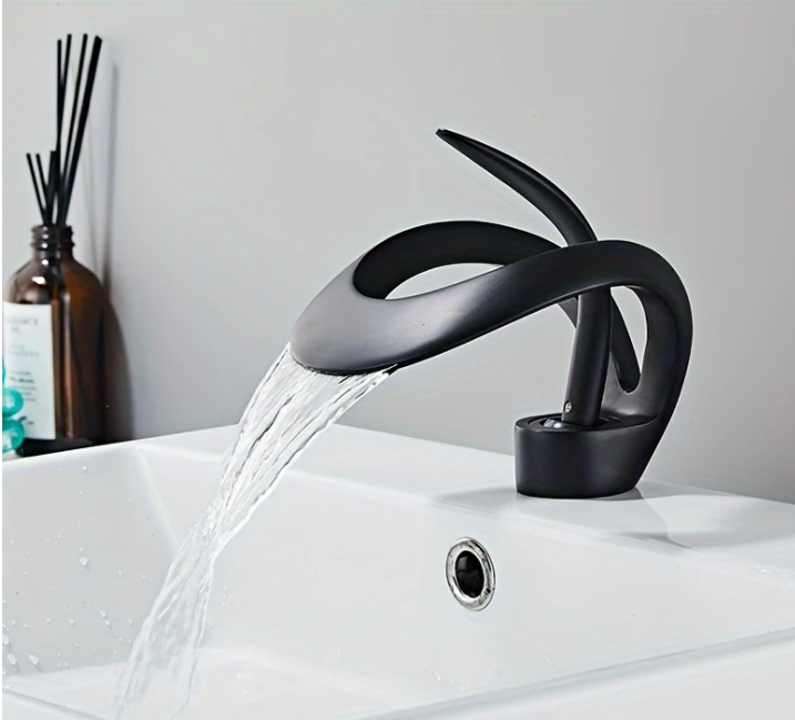 Zwarte wastafelkraan met gebogen sculpturaal ontwerp, gemonteerd op witte wastafel met stromend water; links van de kraan staat een bruine diffuserfles met zwarte geurstokjes en gedeeltelijk zichtbaar etiket; de setting toont een moderne en stijlvolle badkamer met minimalistische uitstraling, een elegante combinatie van functionaliteit en design