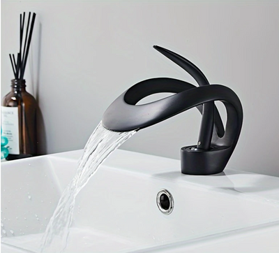 Zwarte wastafelkraan met gebogen sculpturaal ontwerp, gemonteerd op witte wastafel met stromend water; links van de kraan staat een bruine diffuserfles met zwarte geurstokjes en gedeeltelijk zichtbaar etiket; de setting toont een moderne en stijlvolle badkamer met minimalistische uitstraling, een elegante combinatie van functionaliteit en design