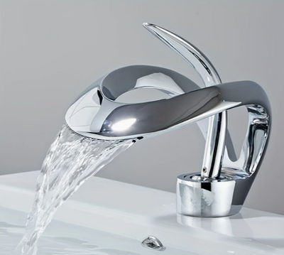 Design wastafelkraan in chroomafwerking met elegant gebogen bladvormige uitloop en bijpassende hendel, gemonteerd op witte wastafel met stromend water; de kraan contrasteert subtiel met de lichtgrijze achterwand, een stijlvolle combinatie van organische vorm en moderne functionaliteit voor hedendaags badkamerdesign