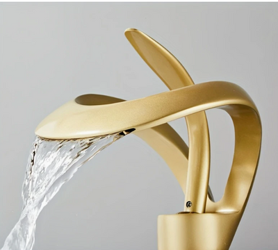 Design wastafelkraan in geborsteld goud met sierlijke gebogen uitloop en geïntegreerde handgreep, gemonteerd op witte wastafel; water stroomt soepel uit de kraan, die zich bevindt in een moderne badkamer met minimalistische uitstraling, een stijlvolle en functionele blikvanger die luxe en elegantie toevoegt aan hedendaags sanitair