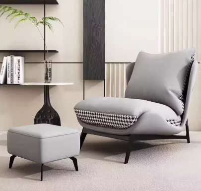 Loungefauteuil met bijpassende ottoman, uitgevoerd in lichtgrijs kunstleer met een pied-de-poule motief op de zijpanelen en achterzijde; het zitmeubel staat op zwarte poten en is geplaatst in een modern interieur met beige wandpanelen, een zwart-witte ronde bijzettafel en decoratieve elementen zoals een glazen vaas met plant en wandplanken met boeken; het geheel straalt stijl en comfort uit en is perfect voor een eigentijdse woonruimte of relaxhoek