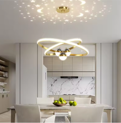 Nordic LED-kroonluchter met gouden interlockende ringen en geïntegreerde LED-bollen, sfeervolle sterren- en maanprojectie, hangend boven een eettafel met schaal groene appels en vier stoelen in een moderne eetkamer met marmeren achterwand