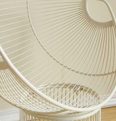 Moderne witte draadstoel met cirkelvormige en spiraalvormige metalen staven, geplaatst op een houten vloer tegen een beige muur. Het ontwerp bestaat uit concentrische ringen en een gebogen frame dat een geometrisch en luchtig silhouet vormt — een artistiek en eigentijds meubelstuk dat comfort en vormgeving combineert.