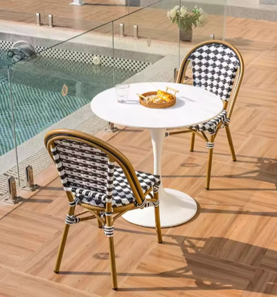 Kleine buitensetting naast een zwembad met ronde witte bistrotafel en twee zwart-wit geweven stoelen met bamboe-look frame en decoratieve bindingsdetails. Op de tafel staan een kom chips, een glas water en een leeg glas. De houten tegelvloer, glazen omheining en bloemen op de achtergrond creëren een elegante en ontspannen sfeer — een charmante plek voor een zomerse pauze in Parijse stijl.
