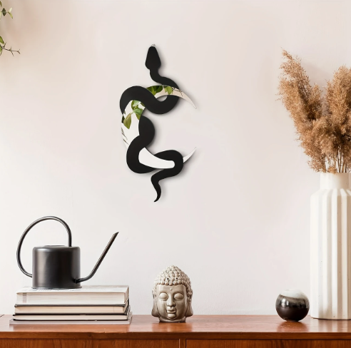 Sfeervolle tabletop-opstelling met decoratieve elementen tegen een witte muur; links een zwarte gieter bovenop drie boeken, centraal een grijzige kandelaar met rustieke uitstraling, en rechts een witte vaas met gedroogde bruine pluimen, vergezeld door een klein rond zwart object; een minimalistisch en natuurlijk interieuraccent dat warmte en textuur toevoegt aan elke ruimte