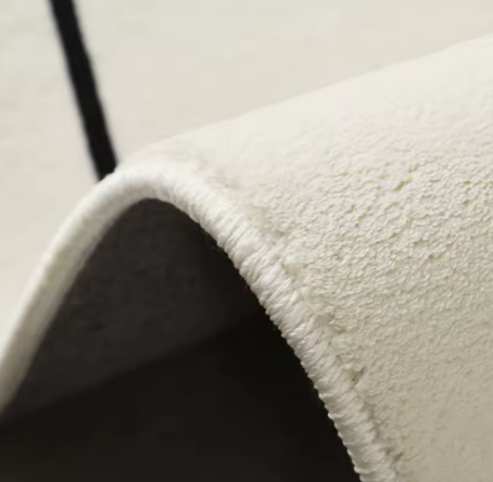 Detailweergave van lichtgekleurd fluffy vloerkleed met hoogpolige textuur, toont gebogen rand met fijne witte stikking en gladde antislip onderzijde; afgebeeld op neutrale ondergrond zonder meubels, een kwaliteitsvol vloerkleed met zachte bovenkant en duurzaam afgewerkte rand voor stijlvol en comfortabel gebruik in modern interieur
