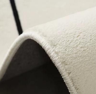 Detailweergave van lichtgekleurd fluffy vloerkleed met hoogpolige textuur, toont gebogen rand met fijne witte stikking en gladde antislip onderzijde; afgebeeld op neutrale ondergrond zonder meubels, een kwaliteitsvol vloerkleed met zachte bovenkant en duurzaam afgewerkte rand voor stijlvol en comfortabel gebruik in modern interieur