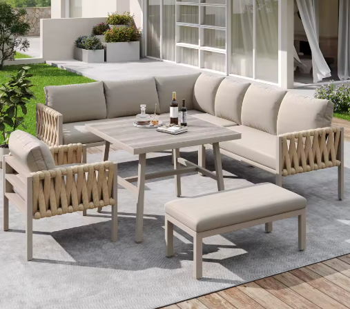Moderne tuinlounge set op een houten terras met groot buitenkleed, bestaande uit een hoekbank, fauteuil, bankje en rechthoekige eettafel met houtlook blad. De meubels zijn afgewerkt met rope-details en lichtbeige kussens. Op de tafel staan wijnflessen, glazen en een dienblad — een elegante setting voor buiten dineren en ontspannen met uitzicht op een strak aangelegde tuin.
