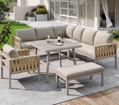 Moderne tuinlounge set op een houten terras met groot buitenkleed, bestaande uit een hoekbank, fauteuil, bankje en rechthoekige eettafel met houtlook blad. De meubels zijn afgewerkt met rope-details en lichtbeige kussens. Op de tafel staan wijnflessen, glazen en een dienblad — een elegante setting voor buiten dineren en ontspannen met uitzicht op een strak aangelegde tuin.